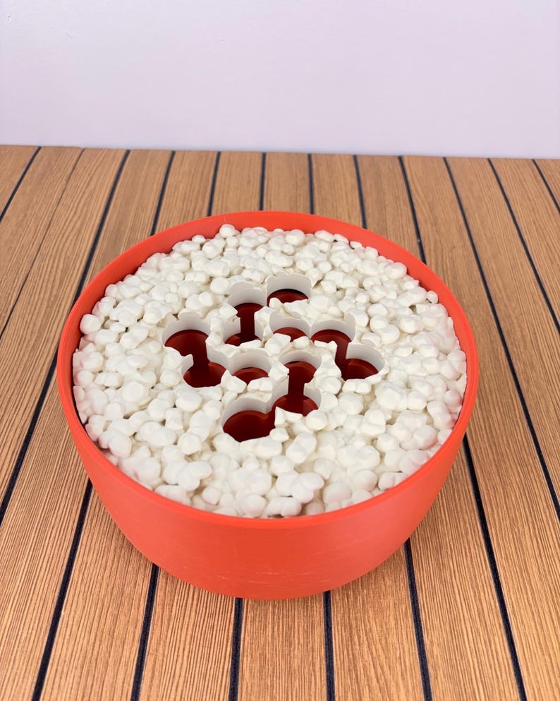 May include: A round, orange bowl filled with small, white, pebble-like objects. The bowl features a central, white, multi-holed insert that reveals a dark red interior, creating a contrast between the white surface and the deep red base.