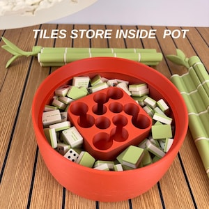 May include: A round orange container filled with small green and white mahjong tiles and a central orange grid insert. The text TILES STORE INSIDE POT is visible above the container, which is flanked by two green bamboo-shaped objects.