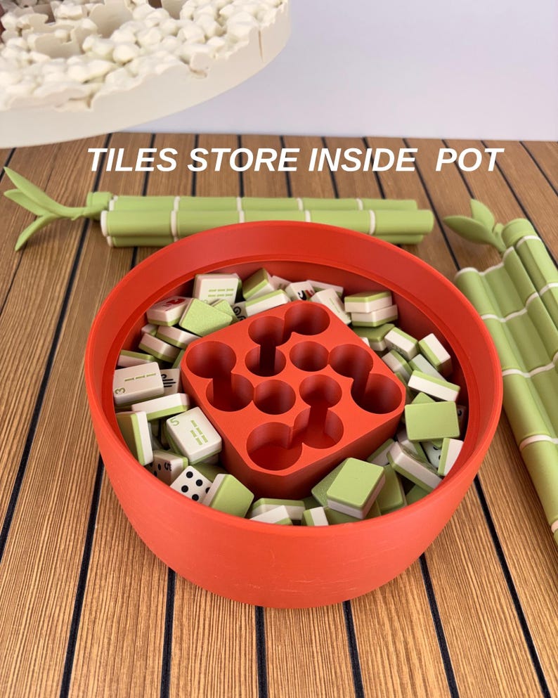 May include: A round orange container filled with small green and white mahjong tiles and a central orange grid insert. The text TILES STORE INSIDE POT is visible above the container, which is flanked by two green bamboo-shaped objects.