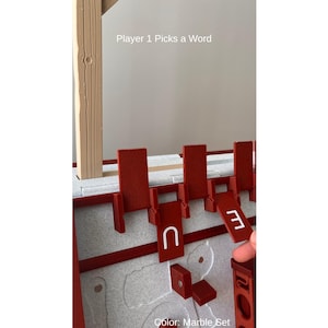 May include: A red and white word game set with the text "Player 1 Picks a Word" and "Color: Marble Set". The game includes red letter holders and letter tiles with the letter "U" and the number "3".