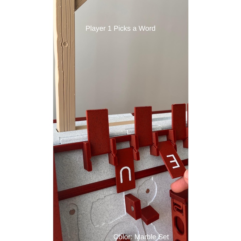 May include: A red and white word game set with the text "Player 1 Picks a Word" and "Color: Marble Set". The game includes red letter holders and letter tiles with the letter "U" and the number "3".