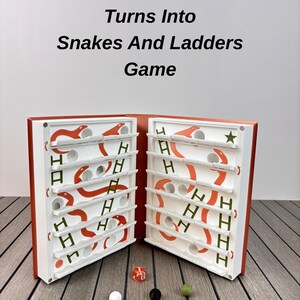 May include: A board game titled Turns Into Snakes And Ladders Game with a copper cover and white interior. The game board features orange snakes, green ladders, and circular holes. Included are one red die and three small spheres in white, black, and green.