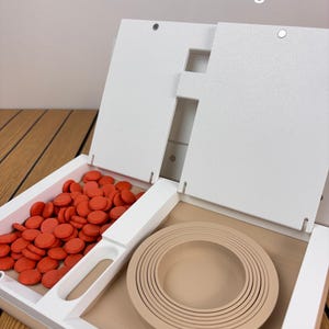 May include: A white storage organizer with the text Pieces Store Inside Pages. The left compartment contains numerous small, round, orange game pieces. The right compartment holds a set of nested, circular, beige rings of varying sizes.