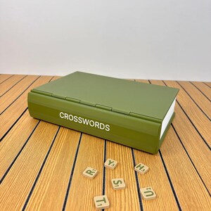 May include: An olive green storage box shaped like a book with the word CROSSWORDS printed in white on the spine. Five small square tiles with letters and numbers are scattered in front of the box, including R1, Y3, T1, S1, H3, and U2.