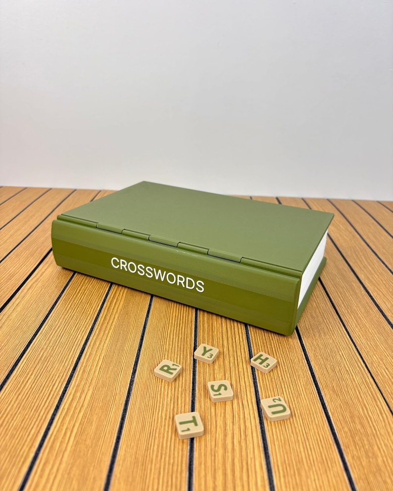 May include: An olive green storage box shaped like a book with the word CROSSWORDS printed in white on the spine. Five small square tiles with letters and numbers are scattered in front of the box, including R1, Y3, T1, S1, H3, and U2.