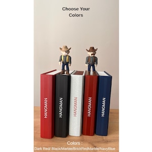 May include: Five colourful bookends with the word "HANGMAN" printed on the spines. Each bookend is a different colour: dark red, black, marble, brick red marble, and navy blue. Two cowboy figurines stand on top. The text "Choose Your Colors" is at the top.