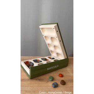 Pode incluir: Um tabuleiro de Mancala aberto em verde-oliva e bege, com pedras coloridas. O tabuleiro est&aacute; sobre uma superf&iacute;cie de madeira, com a palavra "MANCALA" impressa na frente. Cor: Verde-oliva / Bege.