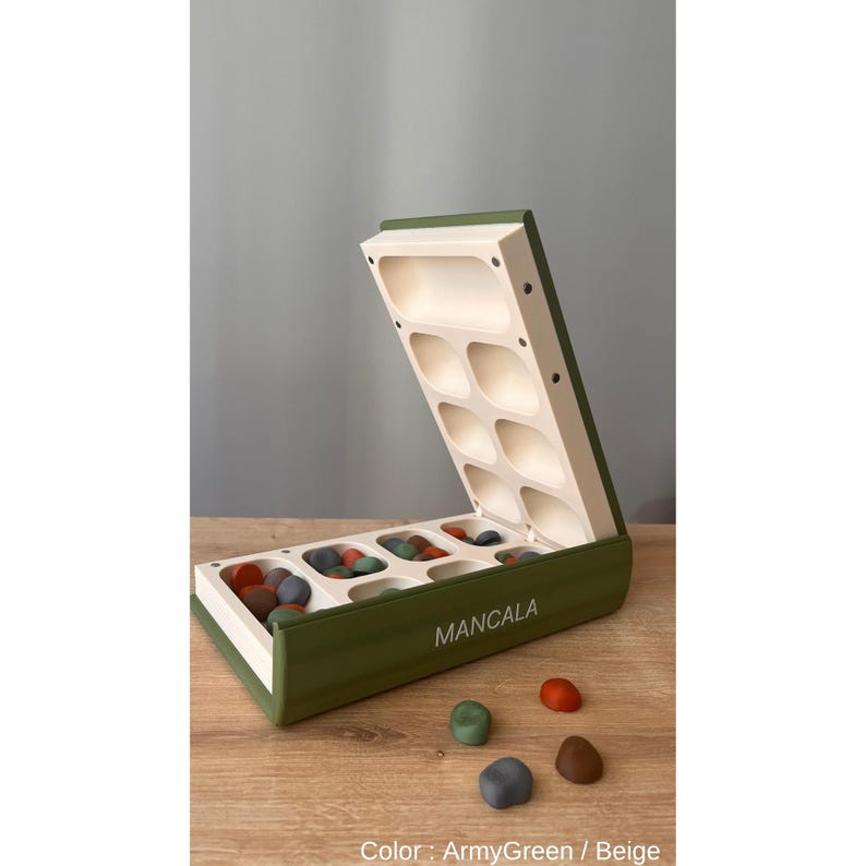 Pode incluir: Um tabuleiro de Mancala aberto em verde-oliva e bege, com pedras coloridas. O tabuleiro est&aacute; sobre uma superf&iacute;cie de madeira, com a palavra "MANCALA" impressa na frente. Cor: Verde-oliva / Bege.