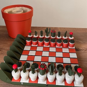 K&ouml;nnte beinhalten: Ein Schachspiel mit einem Brett aus abwechselnd orangefarbenen und wei&szlig;en Quadraten, umrandet von einem dunkelgr&uuml;nen Rahmen. Die Spielfiguren sind kleine gr&uuml;ne Kakteen in T&ouml;pfen, einige mit roten Akzenten, auf dem Brett. Ein gro&szlig;er orangefarbener Topf steht daneben.