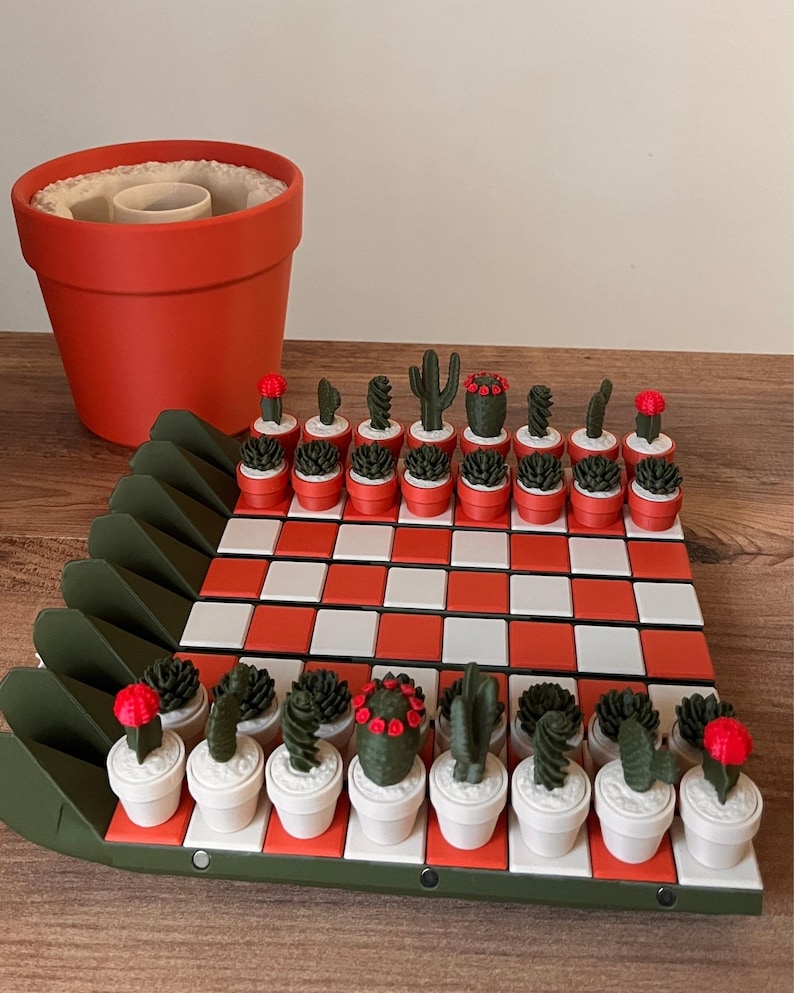 K&ouml;nnte beinhalten: Ein Schachspiel mit einem Brett aus abwechselnd orangefarbenen und wei&szlig;en Quadraten, umrandet von einem dunkelgr&uuml;nen Rahmen. Die Spielfiguren sind kleine gr&uuml;ne Kakteen in T&ouml;pfen, einige mit roten Akzenten, auf dem Brett. Ein gro&szlig;er orangefarbener Topf steht daneben.