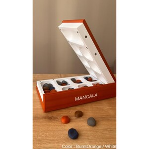 Pode incluir: Um jogo de tabuleiro Mancala laranja queimado e branco com uma tampa articulada. O tabuleiro est&aacute; aberto, revelando compartimentos brancos cheios de pedras coloridas. A palavra "MANCALA" est&aacute; impressa na lateral. V&aacute;rias pedras est&atilde;o espalhadas na superf&iacute;cie de madeira.