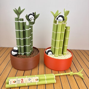 May include: Two decorative planters shaped like bamboo stalks with small panda figures. One planter is brown with dark soil, the other is orange with white pebbles. A separate bamboo stalk piece lies horizontally with mahjong tiles attached.