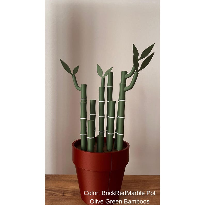 May include: A decorative arrangement of olive green bamboo stalks with white bands and dark green leaves, planted in a brick-red marble pot. The pot sits on a wooden surface against a neutral background.