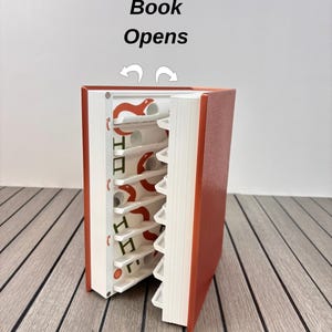 May include: A copper-colored book with a white interior storage system. The book opens to reveal white shelves featuring orange snake graphics and small white circular cutouts. Text above the book reads Book Opens, and text below reads Colors: Copper Cover / White.