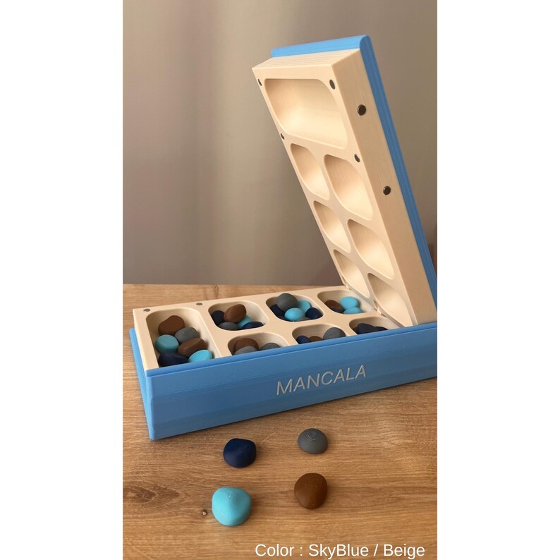 Pode incluir: Um jogo de tabuleiro Mancala azul celeste e bege com tampa articulada. O tabuleiro &eacute; preenchido com pequenas pedras coloridas em tons de azul, castanho e cinza. A palavra "MANCALA" est&aacute; impressa na lateral do tabuleiro. A cor do jogo &eacute; Azul celeste / Bege.