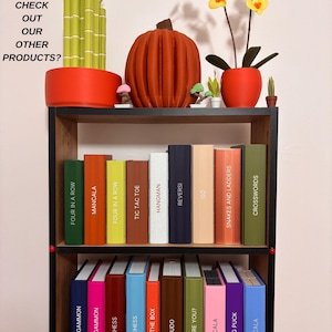 May include: A black bookshelf holding two rows of colorful game boxes labeled with titles like Four in a Row, Mancala, Chess, and Snakes and Ladders. The top shelf features a 3D printed bamboo plant, a pumpkin, and an orchid. Text reads: DID YOU CHECK OUT OUR OTHER PRODUCTS?