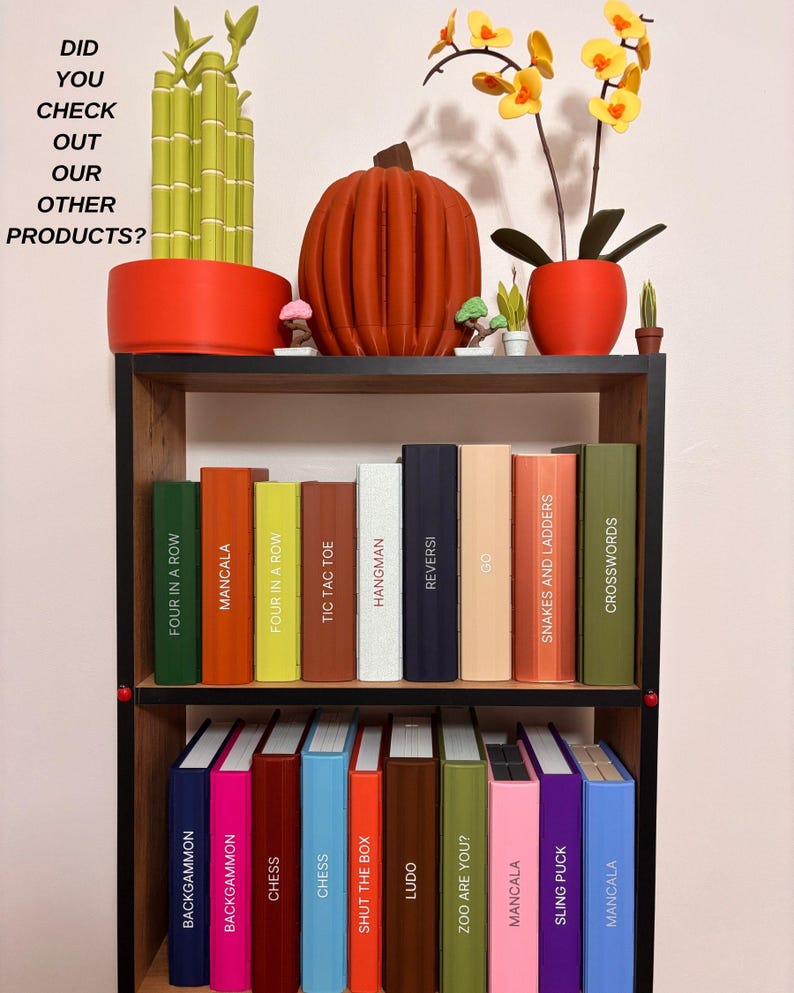 May include: A black bookshelf holding two rows of colorful game boxes labeled with titles like Four in a Row, Mancala, Chess, and Snakes and Ladders. The top shelf features a 3D printed bamboo plant, a pumpkin, and an orchid. Text reads: DID YOU CHECK OUT OUR OTHER PRODUCTS?