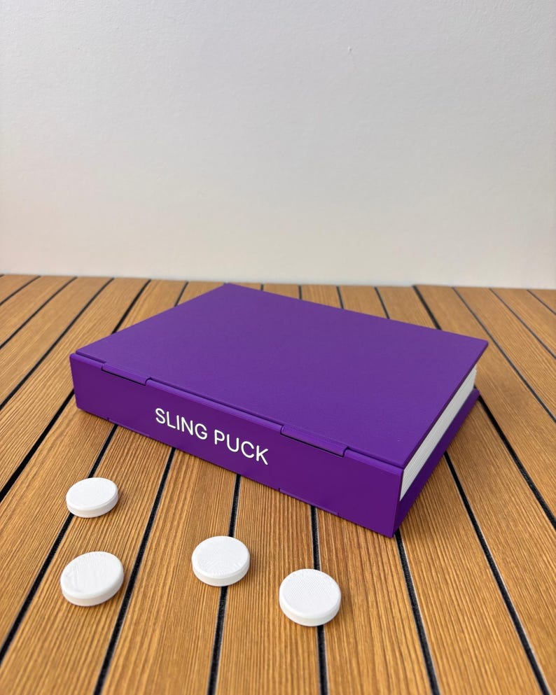 May include: A rectangular purple game board with the text SLING PUCK printed in white on the side. The game board features a hinged design and is accompanied by three small, round, white plastic pucks placed on the surface nearby.