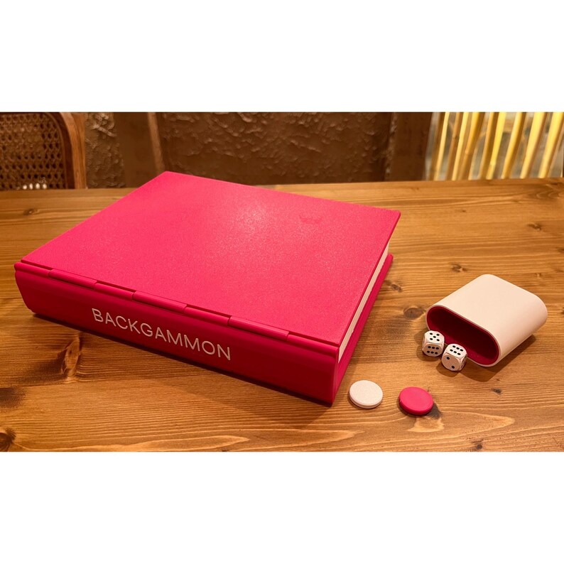 May include: A hot pink backgammon set with the game's name printed in white. The set includes dice, game pieces, and a dice cup. The game is displayed on a wooden surface.
