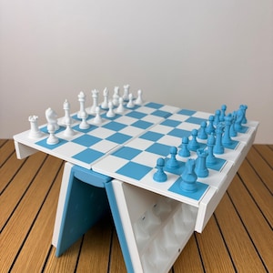 K&ouml;nnte beinhalten: Ein klappbares Schachspiel mit einem Spielbrett im hellblauen und wei&szlig;en Schachbrettmuster. Das Set enth&auml;lt einen vollst&auml;ndigen Satz wei&szlig;er und hellblauer Schachfiguren, die auf dem Brett angeordnet sind. Der Sockel ist wei&szlig; und hellblau.