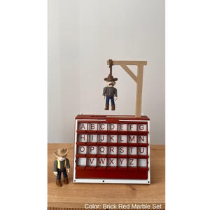 May include: A brick red marble set with a western theme. The set includes a gallows with a cowboy figure hanging from it, and a second cowboy figure standing on the left. The set has a letter board with the alphabet.