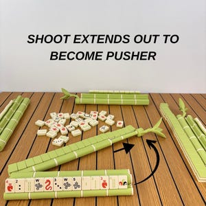 May include: A Mahjong set featuring light green tile racks designed to look like bamboo stalks. The racks include an extendable pusher arm. The text SHOOT EXTENDS OUT TO BECOME PUSHER is visible above the tiles and racks on the surface.