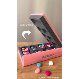Pode incluir: Jogo de tabuleiro Mancala rosa e cinza com pe&ccedil;as coloridas. O tabuleiro aberto revela compartimentos cheios de pedras azuis, rosas e brancas. O texto "Mancala" est&aacute; impresso na lateral.