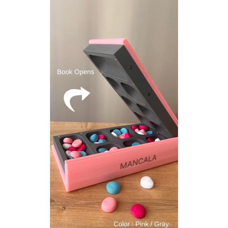 Pode incluir: Jogo de tabuleiro Mancala rosa e cinza com pe&ccedil;as coloridas. O tabuleiro aberto revela compartimentos cheios de pedras azuis, rosas e brancas. O texto "Mancala" est&aacute; impresso na lateral.