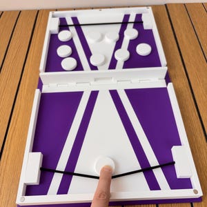 May include: A white and purple board game with the text Lunch Pucks Through Gate. The game features a central gate, two black elastic bands, and several white circular pucks. One puck is positioned against an elastic band on the board.