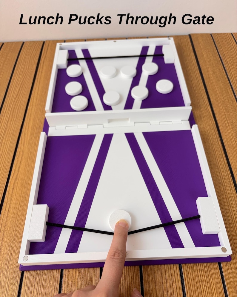 May include: A white and purple board game with the text Lunch Pucks Through Gate. The game features a central gate, two black elastic bands, and several white circular pucks. One puck is positioned against an elastic band on the board.