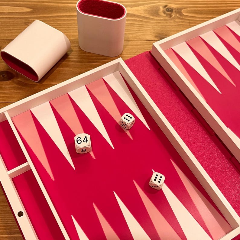 May include: A backgammon set with a vibrant pink playing surface and white and pink triangular points. The set includes two white dice, a dice cup, and a container. The dice show the numbers 64, 8, and 3.