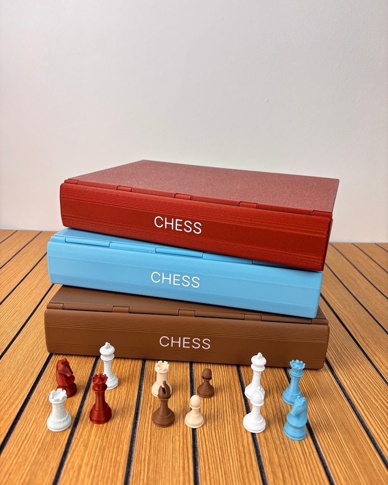 K&ouml;nnte beinhalten: Ein Stapel aus drei rechteckigen Schachspielkoffern in Rot, Hellblau und Braun, wobei auf jeder Seite das Wort CHESS in Wei&szlig; aufgedruckt ist. Eine Sammlung kleiner Schachfiguren in Wei&szlig;, Rot, Braun und Blau ist davor angeordnet.