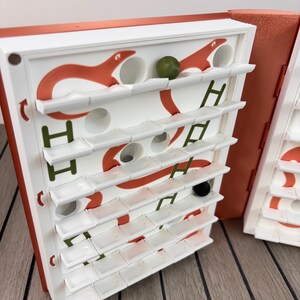 May include: A vertical board game with a white grid featuring orange snake illustrations and green ladder graphics. The game includes small white shelves, a single green marble, a black marble, and a metallic orange die resting on the surface.