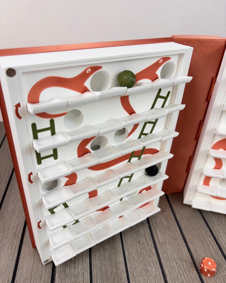 May include: A vertical board game with a white grid featuring orange snake illustrations and green ladder graphics. The game includes small white shelves, a single green marble, a black marble, and a metallic orange die resting on the surface.