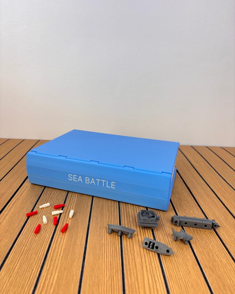 May include: A light blue rectangular board game case with the text SEA BATTLE printed in white on the front. Scattered in front of the case are several small red and white plastic pegs and four miniature gray plastic ship game pieces.