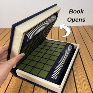 以下が含まれることがあります： 濃い青とクリーム色の本型収納ボックス。開くとオリーブグリーンの正方形のグリッドと、白黒のストライプ模様の円筒形ローラーが2つ現れます。白い矢印と共に「Book Opens」というテキストが印刷されています。