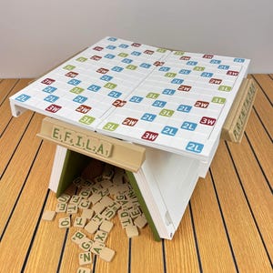 May include: A white word game board with a grid of colored squares labeled 2L, 3L, 2W, and 3W, supported by a tan structure. A tan tile rack displays the letters E, F, I, L, 2, A, and a pile of scattered tan letter tiles sits underneath the board.