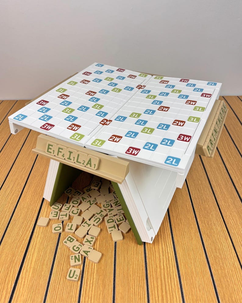 May include: A white word game board with a grid of colored squares labeled 2L, 3L, 2W, and 3W, supported by a tan structure. A tan tile rack displays the letters E, F, I, L, 2, A, and a pile of scattered tan letter tiles sits underneath the board.