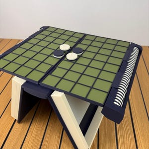 以下が含まれることがあります： Turns Into Reversi Gameというテキストが書かれた折りたたみ式のボードゲーム。ダークブルーのフレーム、オリーブグリーンのグリッド面、白とダークブルーの円形の駒が特徴です。構造はオフホワイトの斜めの脚で支えられています。