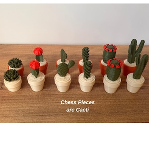K&ouml;nnte beinhalten: Eine Sammlung von Miniatur-Schachfiguren in Form verschiedener gr&uuml;ner Kakteen in kleinen beigen und orangefarbenen T&ouml;pfen. Einige Kakteen haben leuchtend rote Bl&uuml;ten. Der Text "Chess Pieces are Cacti" ist in Wei&szlig; auf die Holzoberfl&auml;che gedruckt.