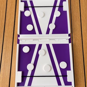 May include: A rectangular wooden game board featuring a white and purple geometric design with black elastic bands at each end. The board includes several white circular game pieces and displays the text Turns Into Sling Puck at the top.