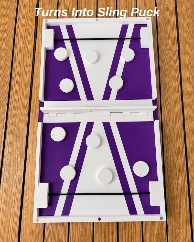 May include: A rectangular wooden game board featuring a white and purple geometric design with black elastic bands at each end. The board includes several white circular game pieces and displays the text Turns Into Sling Puck at the top.