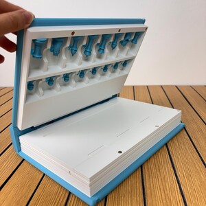 K&ouml;nnte beinhalten: Ein hellblaues, buchf&ouml;rmiges Aufbewahrungsetui mit wei&szlig;em Innenraum. Der obere Teil verf&uuml;gt &uuml;ber geformte Schlitze, die kleine blaue Schachfiguren halten. Der untere Teil ist eine flache wei&szlig;e Oberfl&auml;che mit magnetischen Verschlusspunkten.