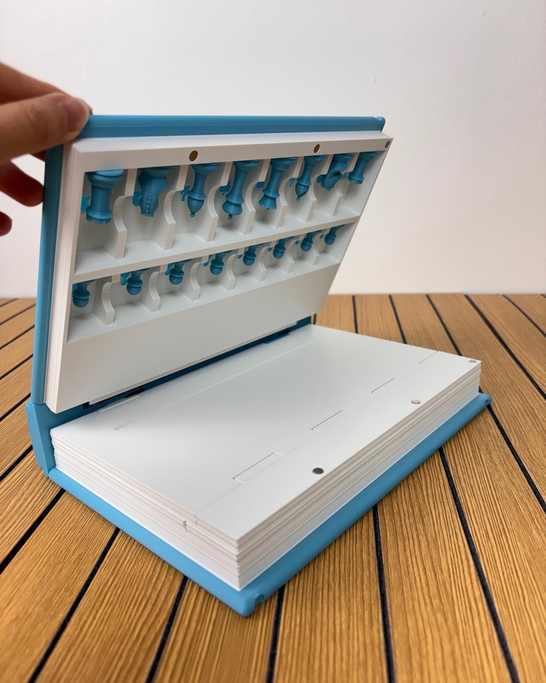 K&ouml;nnte beinhalten: Ein hellblaues, buchf&ouml;rmiges Aufbewahrungsetui mit wei&szlig;em Innenraum. Der obere Teil verf&uuml;gt &uuml;ber geformte Schlitze, die kleine blaue Schachfiguren halten. Der untere Teil ist eine flache wei&szlig;e Oberfl&auml;che mit magnetischen Verschlusspunkten.