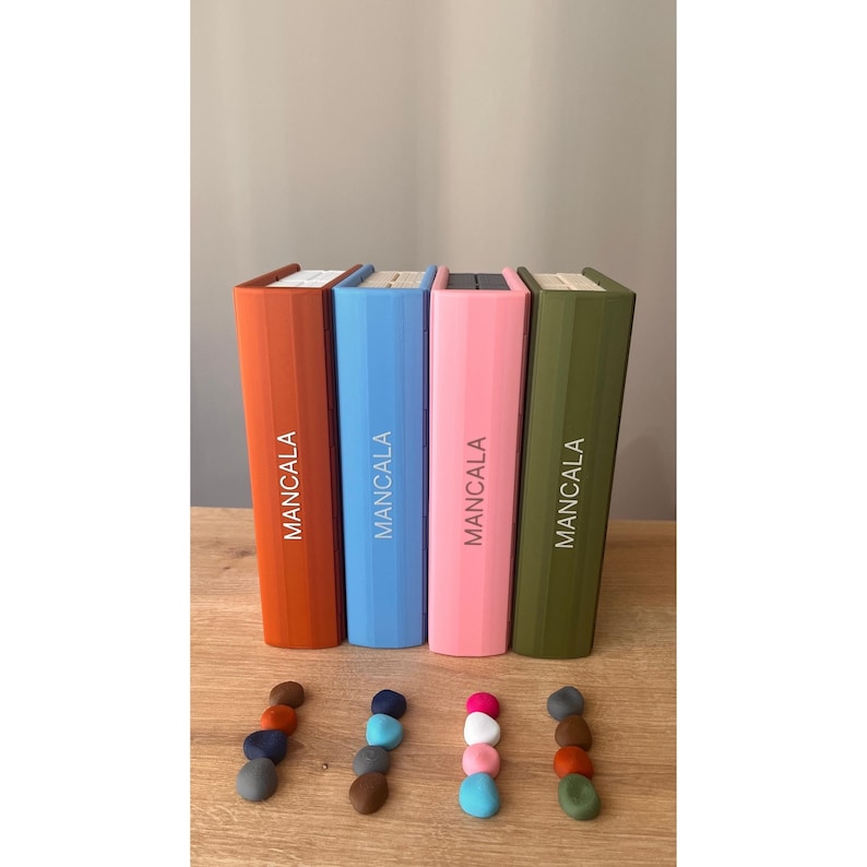 Pode incluir: Quatro caixas de jogo Mancala coloridas, laranja, azul, rosa e verde-oliva, est&atilde;o em p&eacute; sobre uma superf&iacute;cie de madeira. Abaixo, pequenas pilhas de pe&ccedil;as coloridas do jogo est&atilde;o dispostas. A palavra "MANCALA" est&aacute; impressa verticalmente em cada caixa.