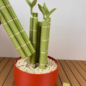 May include: A decorative bamboo-style plant sculpture featuring segmented light green stalks with white rings, set in a solid orange cylindrical pot filled with small white pebbles. A separate small stack of green and white segments sits nearby.
