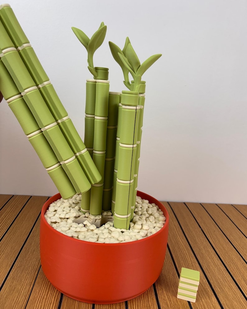 May include: A decorative bamboo-style plant sculpture featuring segmented light green stalks with white rings, set in a solid orange cylindrical pot filled with small white pebbles. A separate small stack of green and white segments sits nearby.