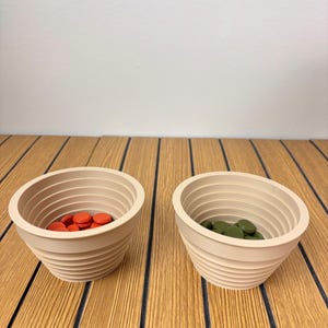 May include: Two beige, ribbed, conical bowls. The left bowl contains small, round, orange discs, while the right bowl contains small, round, olive green discs. Both bowls feature a series of horizontal ridges along their interior and exterior walls.