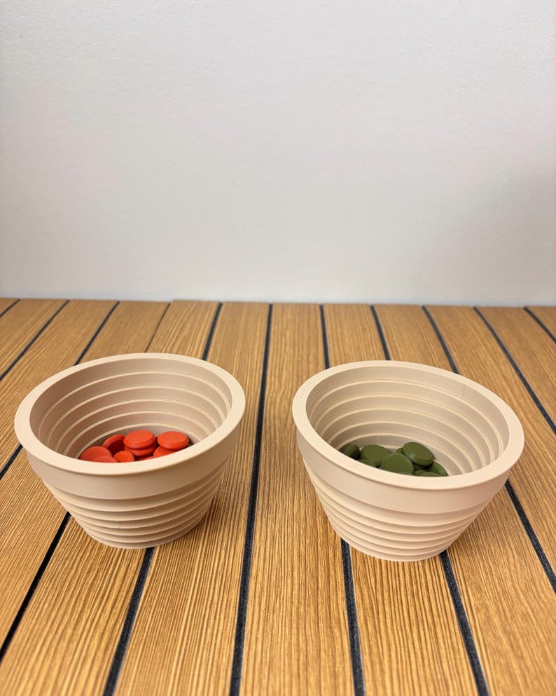May include: Two beige, ribbed, conical bowls. The left bowl contains small, round, orange discs, while the right bowl contains small, round, olive green discs. Both bowls feature a series of horizontal ridges along their interior and exterior walls.