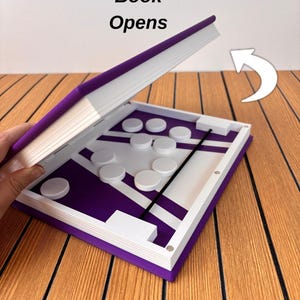 May include: A purple and white game board shaped like a book with the text Book Opens. The interior features a purple surface with white lines, white circular game pieces, and a black elastic band. A white arrow indicates the opening direction.